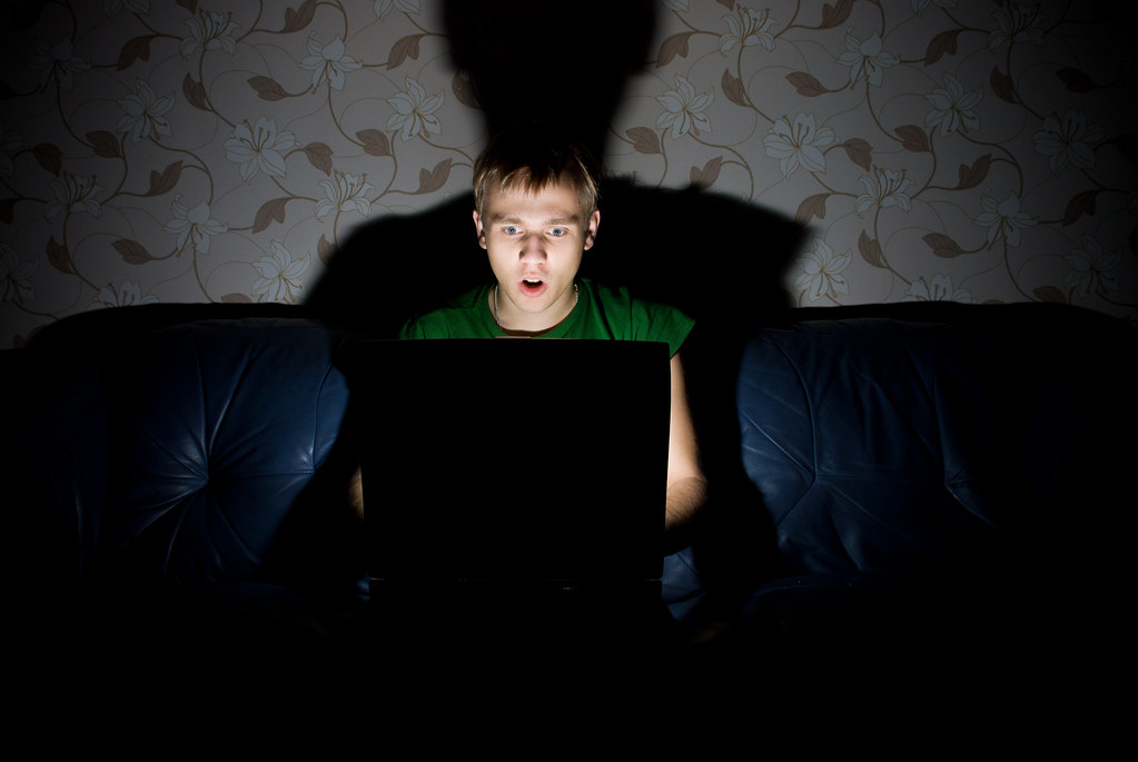 A young man staring at his screen in horror, in a dark room.