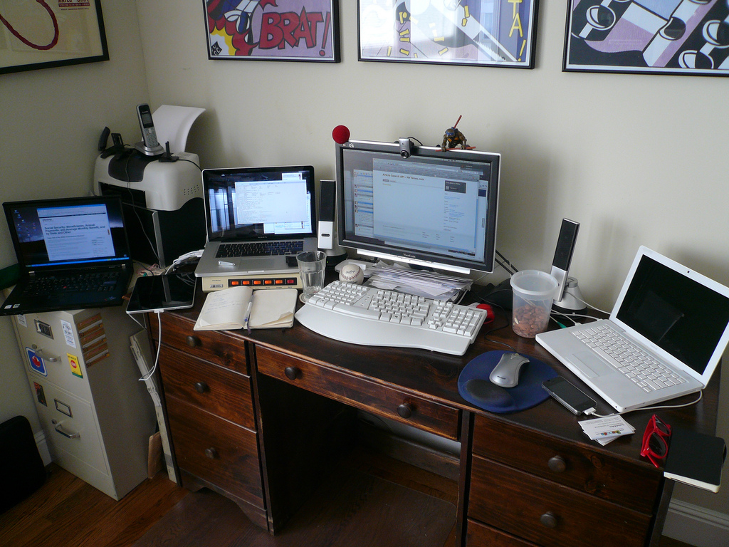 Several computers, a tablet, and a cellphone placed on a relatively small desk.
